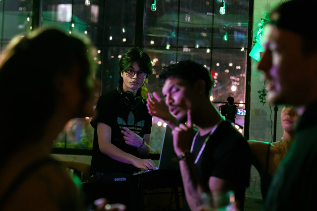 A DJ with headphones is performing at a lively event, surrounded by partygoers who are engaged in conversation. The atmosphere is vibrant with string lights and a large window showing city lights outside.