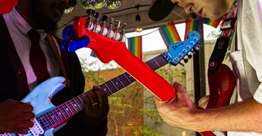 Close-up of two musicians playing colorful electric guitars, one in red and one in blue, at a performance with rainbow decorations in the background.