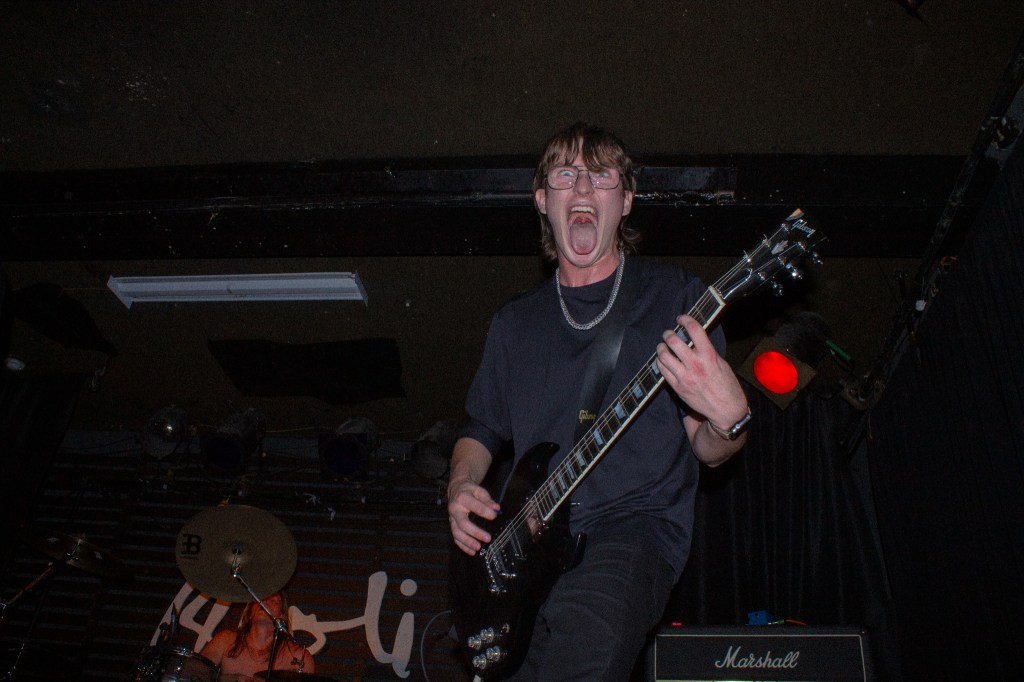 A guitarist performing energetically on stage, shouting into the microphone, with a drummer visible in the background.