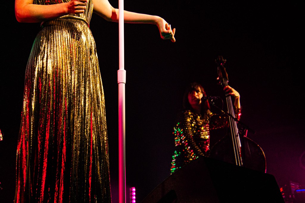 A performer in a shimmering gold dress gestures with one hand on stage, while a musician in a colorful sequined outfit plays the double bass in the background under bright stage lights.