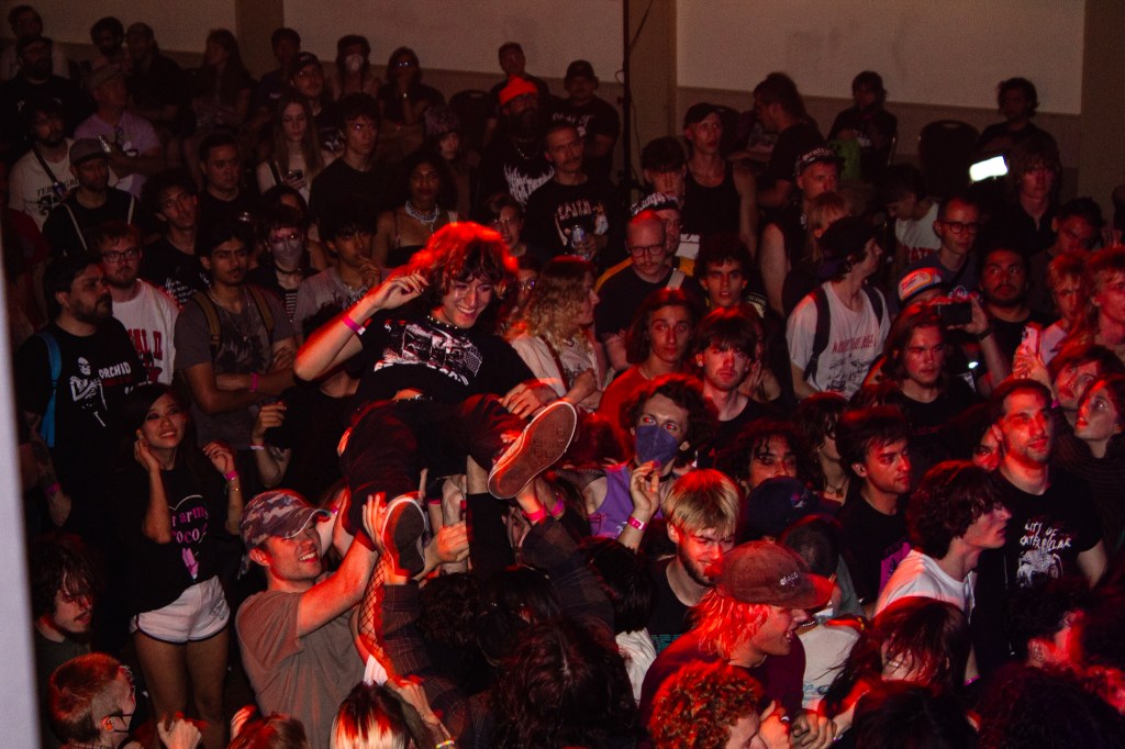 A lively crowd at a concert, with a person crowd surfing above the audience. Various attendees are enjoying the show, with a mix of expressions and styles.