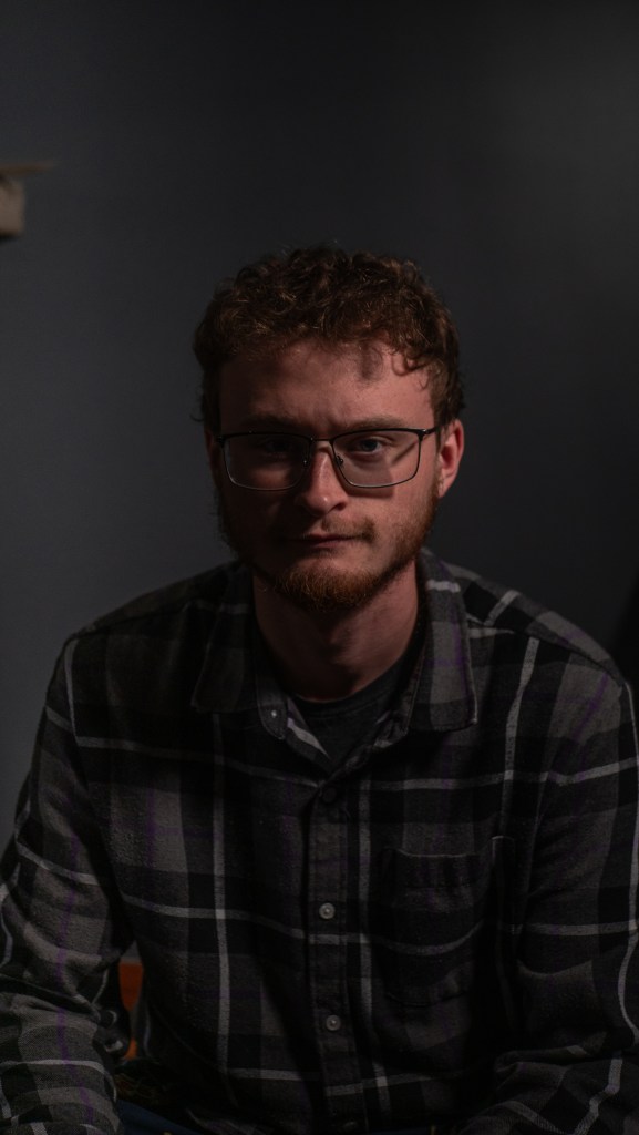 A young man with curly hair and glasses, wearing a plaid shirt, sitting against a dark background.