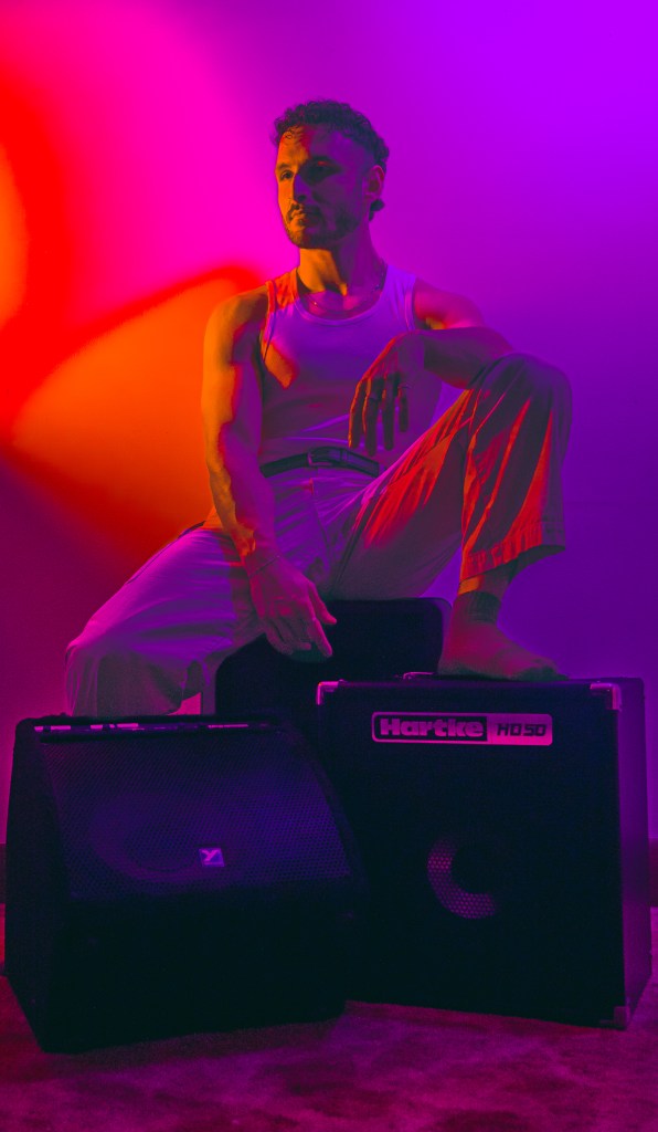 A man with curly hair poses next to black speaker cabinets against a colorful backdrop of purple and orange lighting, wearing a sleeveless top and light-colored pants.