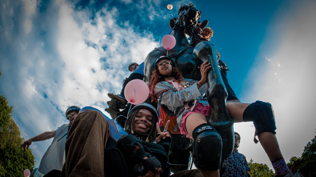 A group of young people posing playfully around a large statue, with some holding pink balloons, set against a blue sky with scattered clouds.