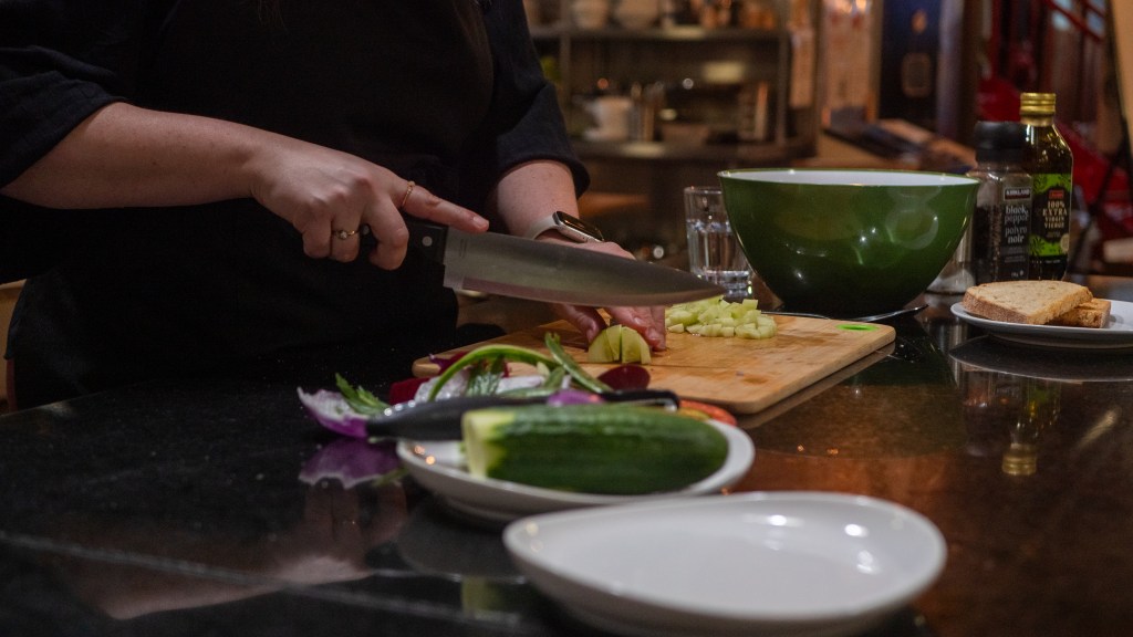 A person chopping vegetables on a wooden cutting board, with a knife in hand. In the background, a green mixing bowl and various ingredients including cucumber, herbs, and slices of bread are visible.