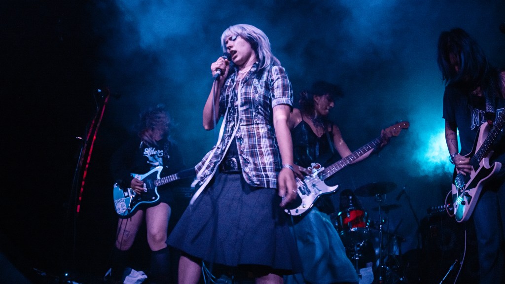 A female lead singer performing on stage with a microphone, surrounded by three bandmates playing electric guitars, against a smoky backdrop with blue lighting.