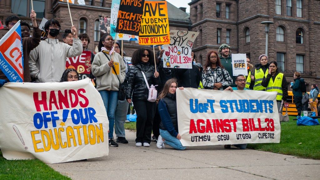 A group of students and activists protesting against Bill 33, holding banners that say 'Hands Off Our Education' and 'UofT Students Against Bill 33' at a rally in front of a government building.